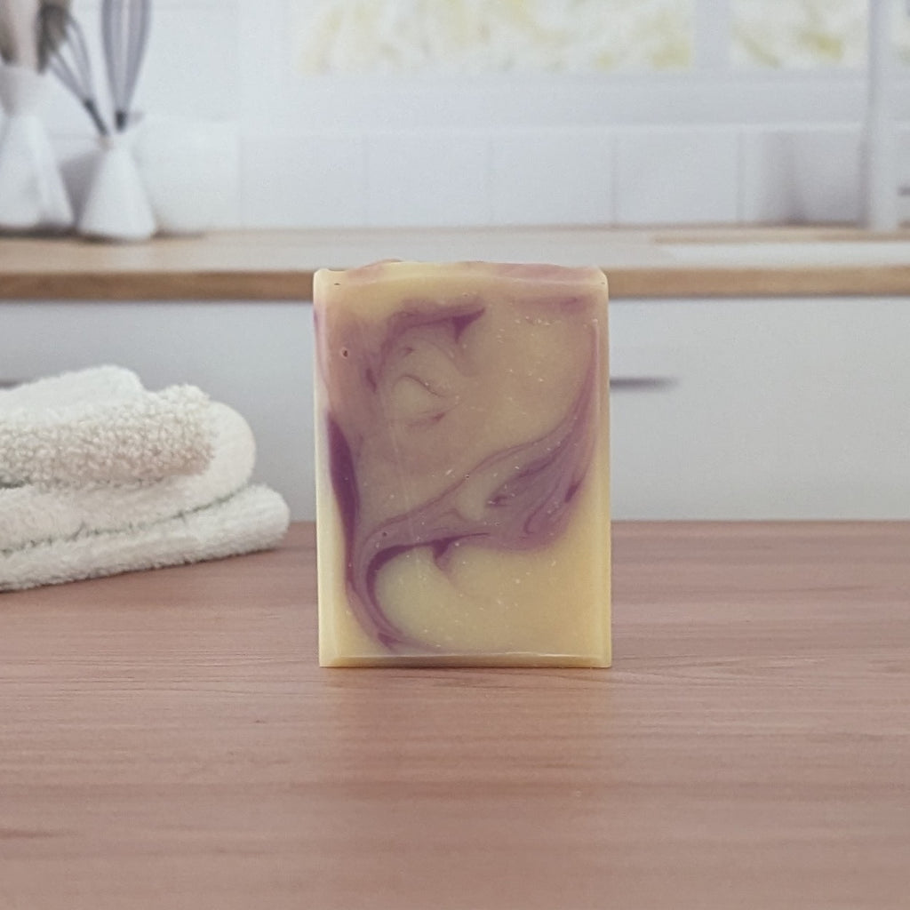 A bar of yellow and purple swirled soap on a light wood surface with a white towel and window in the background.