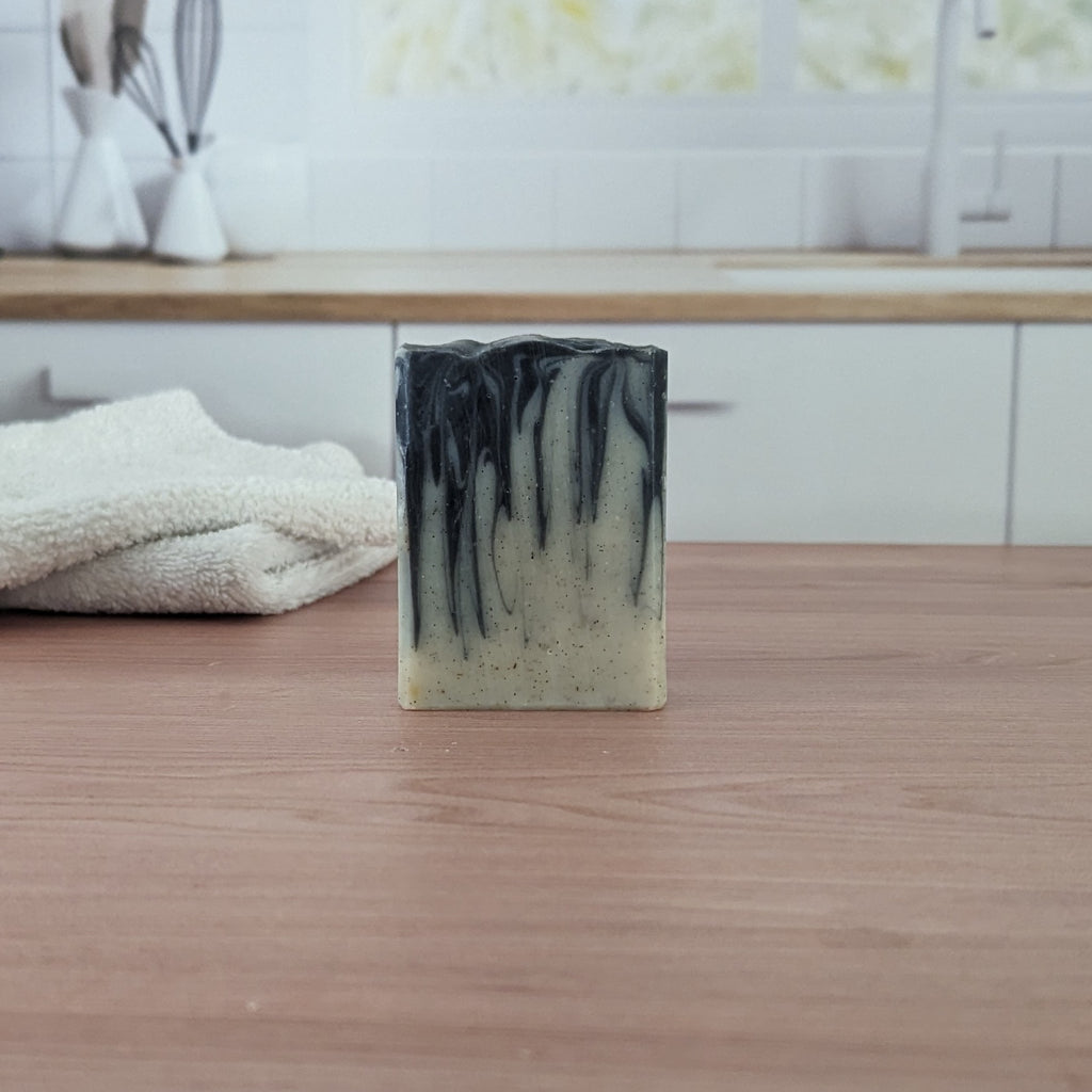 A bar of soap with black dragged into an off-white bottom with flecks on a light wood surface and with a white towel and window.